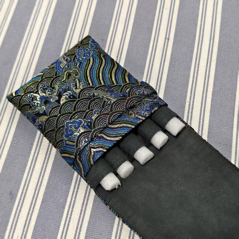 May include: A black pen case with a decorative flap featuring a blue, gold, and black wave pattern. The case holds several black cylindrical objects with white foam inserts, likely for protection. The case is on a striped background.