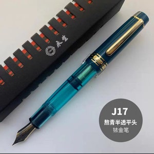 May include: A teal blue fountain pen with a gold trim and a clear ink reservoir. The pen is in a black box with a red and white pattern and the text "J17". The text "J17". The text "款青半透明平头". The text "钛金笔" is also visible.
