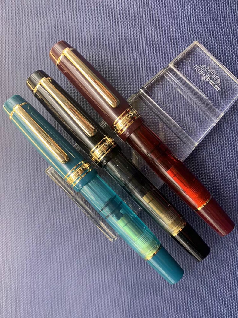 May include: Three fountain pens in a clear acrylic pen holder. The pens are a dark red, black, and teal blue. The pens have gold accents and are all in a horizontal position.