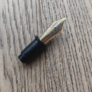 May include: Close-up of a gold fountain pen nib with detailed patterns. The nib is connected to a black pen section. The background is a wooden surface.