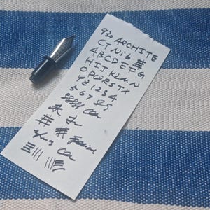 以下が含まれることがあります： 銀色のペン先が付いた黒い万年筆が、手書きの文字が書かれた白い紙の隣に置かれています。テキストには文字、数字、記号が含まれており、「ARCHITE」という単語が見えます。背景は青と白のストライプの生地です。