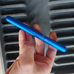 Può includere: Una penna blu metallizzata con la scritta "6u" stampata sul lato. La penna è tenuta in mano su uno sfondo grigio. La penna ha un design elegante e moderno e un cappuccio che sembra essere rimovibile.