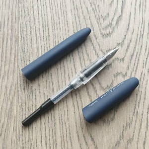May include: A blue fountain pen with a clear barrel and a black nib. The pen is next to a blue pen cap with the word "MINIMALISM" printed on it.