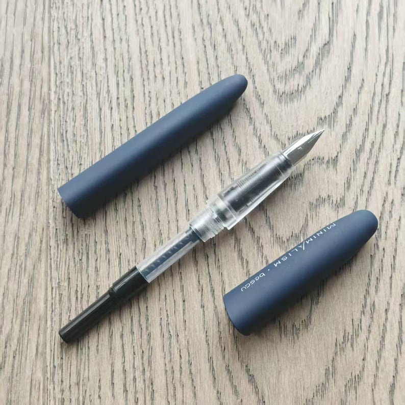 May include: A blue fountain pen with a clear barrel and a black nib. The pen is next to a blue pen cap with the word "MINIMALISM" printed on it.