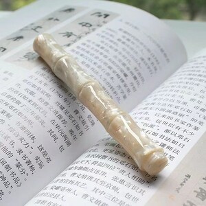 May include: A white and cream colored pen with a textured, marbled finish. The pen is lying on a page of a book with Chinese characters printed on it.