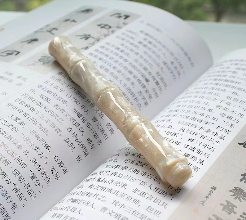 May include: A white and cream colored pen with a textured, marbled finish. The pen is lying on a page of a book with Chinese characters printed on it.