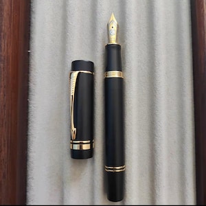 Puede incluir: Una pluma estilogr&aacute;fica negra con detalles dorados y un capuch&oacute;n negro. La pluma tiene una plumilla fina y est&aacute; en posici&oacute;n cerrada.