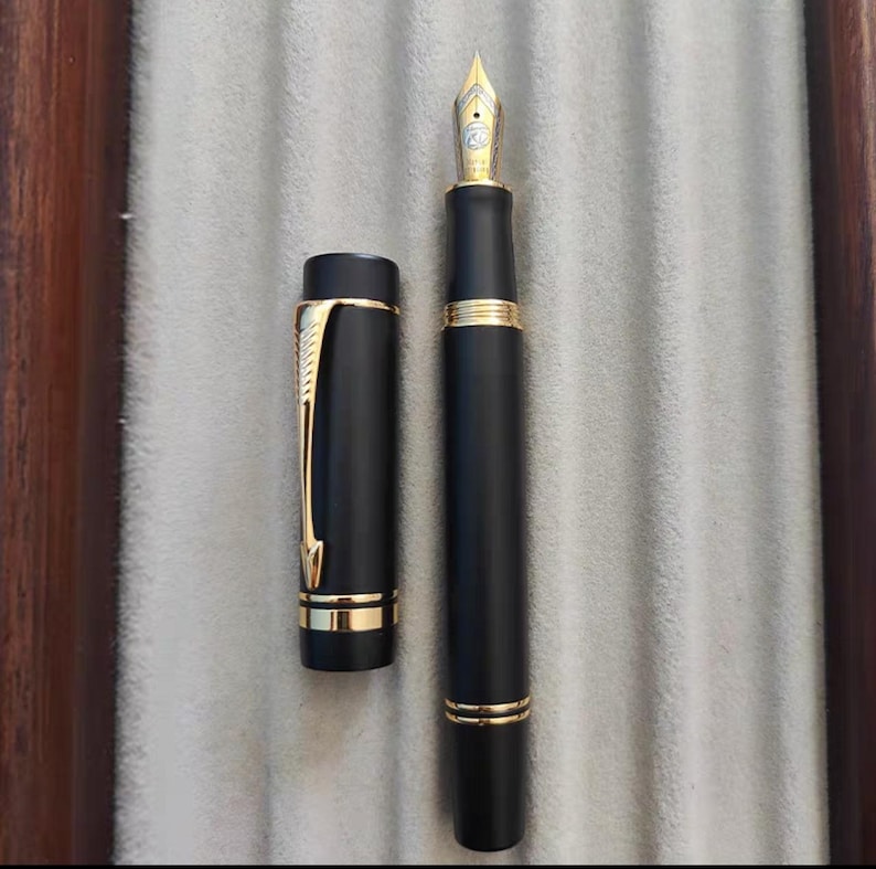 Puede incluir: Una pluma estilogr&aacute;fica negra con detalles dorados y un capuch&oacute;n negro. La pluma tiene una plumilla fina y est&aacute; en posici&oacute;n cerrada.