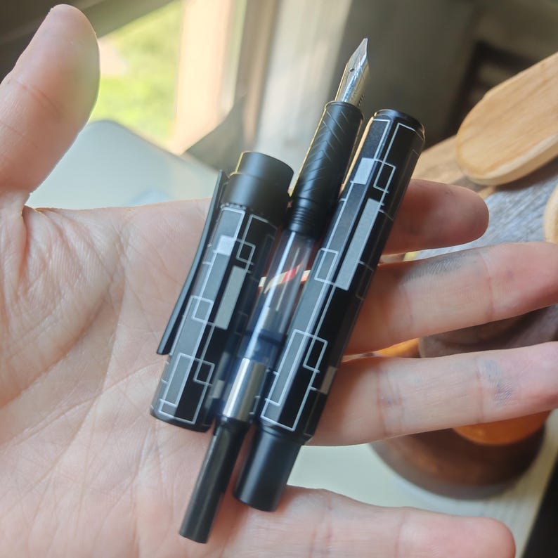 May include: A black fountain pen with a silver nib and a geometric design of white rectangles. The pen is held in a hand, showcasing its sleek design and fine craftsmanship. The pen's cap is also visible.