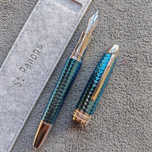 May include: A luxury fountain pen with a blue and green diamond pattern, silver accents, and a gold-colored nib. The pen is next to a gray pen sleeve with the text "St Penpps".