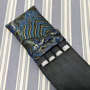 May include: A black pen case with a blue, gold, and black patterned top. The case has a flap that folds over to secure the contents. The interior has four compartments with white foam inserts.