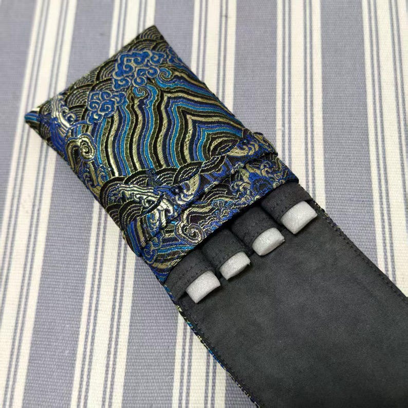 May include: A black pen case with a blue, gold, and black patterned top. The case has a flap that folds over to secure the contents. The interior has four compartments with white foam inserts.
