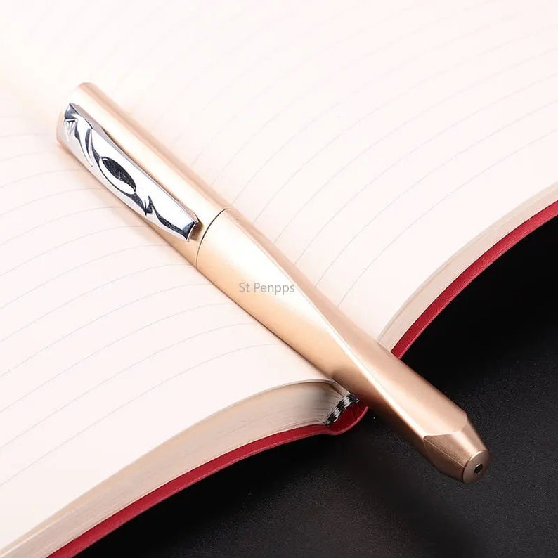 May include: A gold-colored fountain pen with a silver-colored clip and a decorative design on the cap. The pen is lying on a notebook with lined pages.