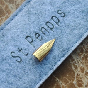 May include: A gold-colored fountain pen nib with a fine point, resting on a light blue felt case. The case has the text "St. Penpps" printed in dark gray. The nib has an engraved design.
