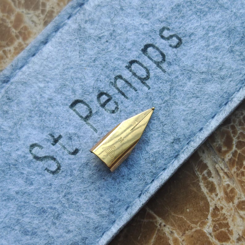 May include: A gold-colored fountain pen nib with a fine point, resting on a light blue felt case. The case has the text "St. Penpps" printed in dark gray. The nib has an engraved design.