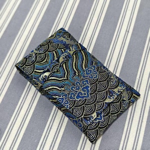 May include: A rectangular, folded fabric pouch with a detailed wave pattern in black, blue, and gold. The pouch is closed and appears to be made of a textured material, possibly silk or a similar fabric. The background is a striped fabric.
