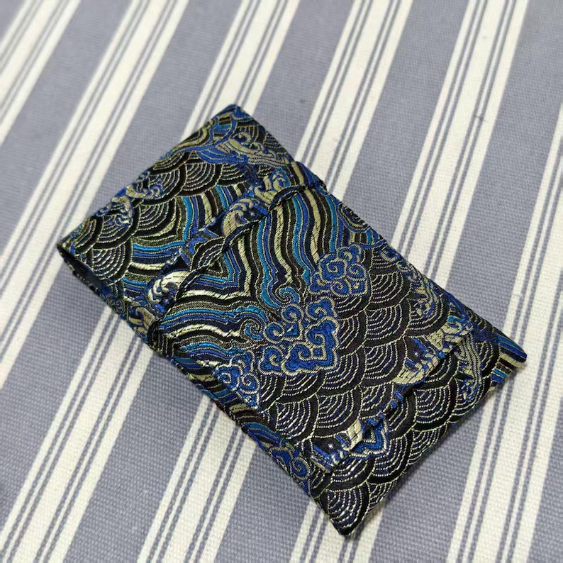 May include: A rectangular, folded fabric pouch with a detailed wave pattern in black, blue, and gold. The pouch is closed and appears to be made of a textured material, possibly silk or a similar fabric. The background is a striped fabric.