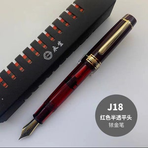 May include: A red and gold fountain pen with a clear ink reservoir. The pen is in a black box with a red and white pattern. The text "J18 红色半透明头 钛金笔" is visible in the image.