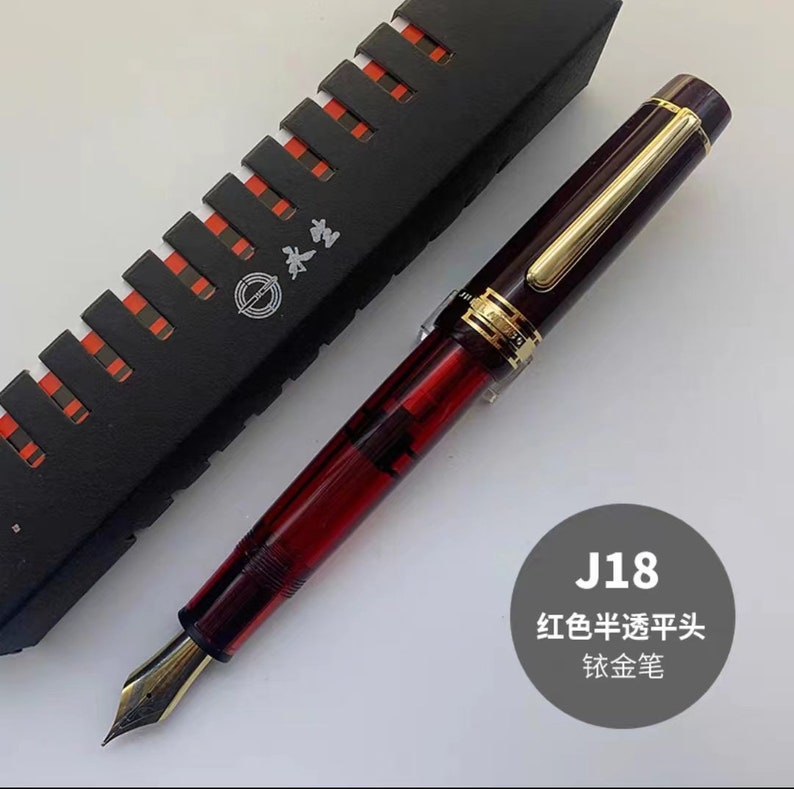 May include: A red and gold fountain pen with a clear ink reservoir. The pen is in a black box with a red and white pattern. The text "J18 红色半透明头 钛金笔" is visible in the image.