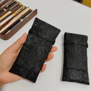 以下が含まれることがあります： テクスチャパターンが施された黒い布製のペンケースが2つ。ケースはフラップ式で、柔らかく高級感のある素材で作られています。