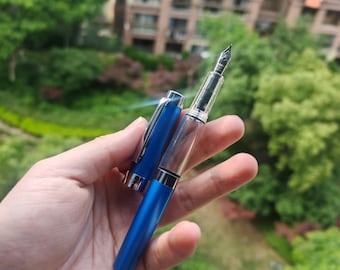 May include: A blue fountain pen with a clear barrel and silver accents. The pen is held in a hand against a blurred background of green foliage and buildings. The pen's cap is open, revealing the nib.