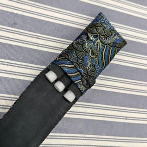 May include: A black and blue pen case with a decorative flap. The flap features a blue, gold, and black wave pattern. The case has three white foam inserts and is set against a striped background.
