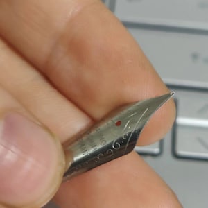 Puede incluir: Primer plano de un plum&iacute;n de pluma estilogr&aacute;fica plateada. El plum&iacute;n est&aacute; sujeto por una mano, con la punta enfocada. El plum&iacute;n tiene marcas grabadas y un peque&ntilde;o punto rojo. El fondo es un teclado borroso.