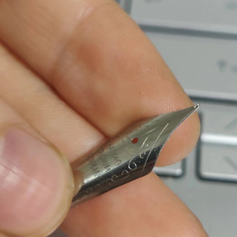 Puede incluir: Primer plano de un plum&iacute;n de pluma estilogr&aacute;fica plateada. El plum&iacute;n est&aacute; sujeto por una mano, con la punta enfocada. El plum&iacute;n tiene marcas grabadas y un peque&ntilde;o punto rojo. El fondo es un teclado borroso.