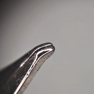 Puede incluir: Primer plano de un objeto met&aacute;lico plateado con una punta redondeada. El objeto tiene una superficie texturizada y parece estar hecho de un material brillante y reflectante. El fondo es un gris suave y borroso.