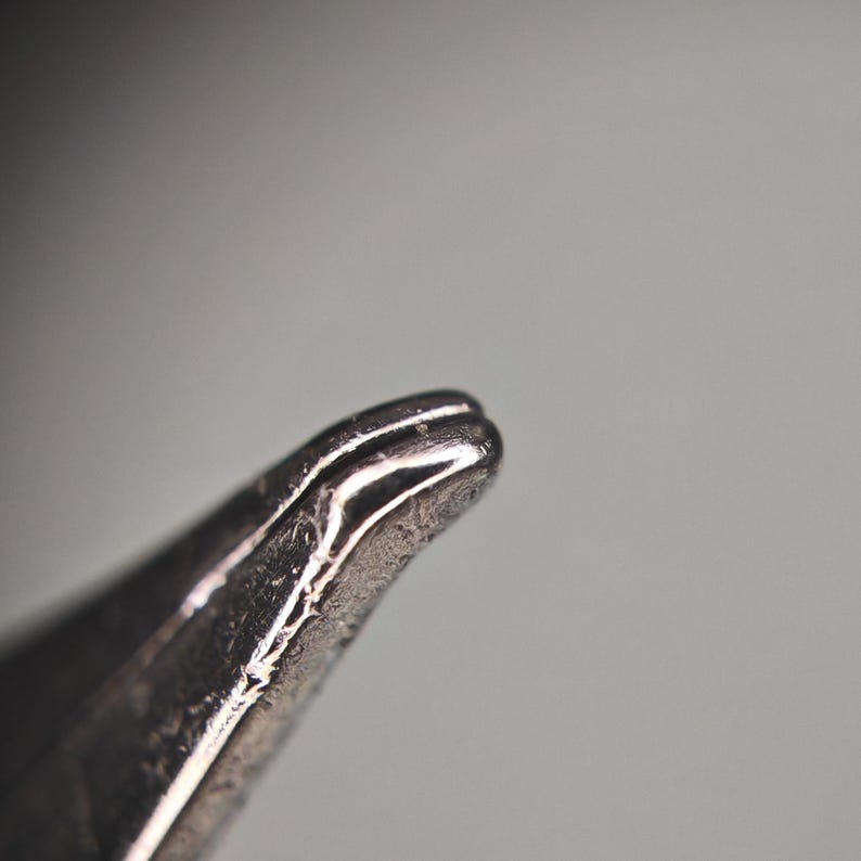 Puede incluir: Primer plano de un objeto met&aacute;lico plateado con una punta redondeada. El objeto tiene una superficie texturizada y parece estar hecho de un material brillante y reflectante. El fondo es un gris suave y borroso.