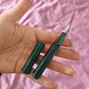 Op de afbeelding: Een groene vulpen met een zilveren penpunt en een bijpassende dop. De pen heeft een gespikkelde, glinsterende afwerking. De dop is losgemaakt, waardoor de penpunt en de inktkamer zichtbaar zijn. De pen wordt in een hand gehouden tegen een roze achtergrond.