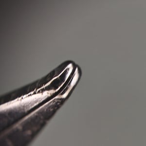 Puede incluir: Primer plano de un objeto met&aacute;lico plateado con una punta curva y afilada. El objeto tiene una superficie reflectante con l&iacute;neas visibles y una ligera hendidura en el extremo. El fondo es gris borroso.