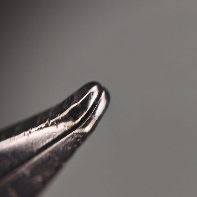 Puede incluir: Primer plano de un objeto met&aacute;lico plateado con una punta curva y afilada. El objeto tiene una superficie reflectante con l&iacute;neas visibles y una ligera hendidura en el extremo. El fondo es gris borroso.