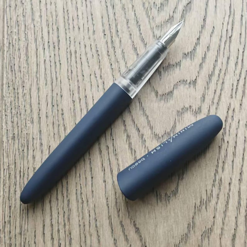 May include: A dark blue fountain pen with a clear plastic barrel and a black cap. The cap has the word "MINIMALISM" engraved on it.
