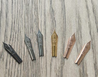 Könnte beinhalten: Sechs Federhalter in verschiedenen Metallfarben, darunter Schwarz, Silber und Gold. Jede Feder hat ein detailliertes Design und die Jahreszahl "2024". Die Federn liegen auf einer Holzoberfläche.