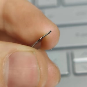 Puede incluir: Primer plano de un objeto met&aacute;lico curvo de color plateado. El objeto tiene una punta redondeada y parece delgado y flexible. El objeto se sostiene entre dos dedos, con un fondo borroso.