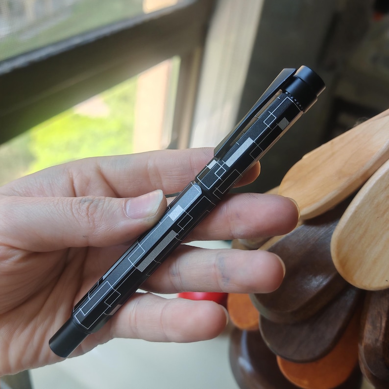 May include: A black pen with a geometric pattern of white and gray rectangles and lines. The pen has a black clip and tip. The pen is held in a hand.