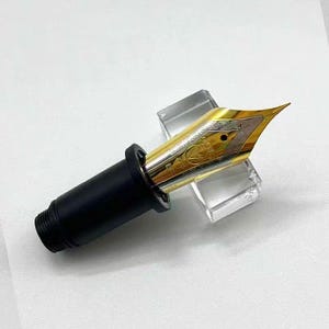 Puede incluir: Primer plano de un plumín de pluma estilográfica con punta dorada y cuerpo negro. El plumín descansa sobre un soporte transparente y rectangular. El plumín dorado tiene diseños grabados intrincados.