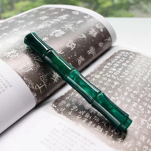 May include: A green fountain pen with a bamboo-like design. The pen is lying on a book with Chinese characters printed on the pages.