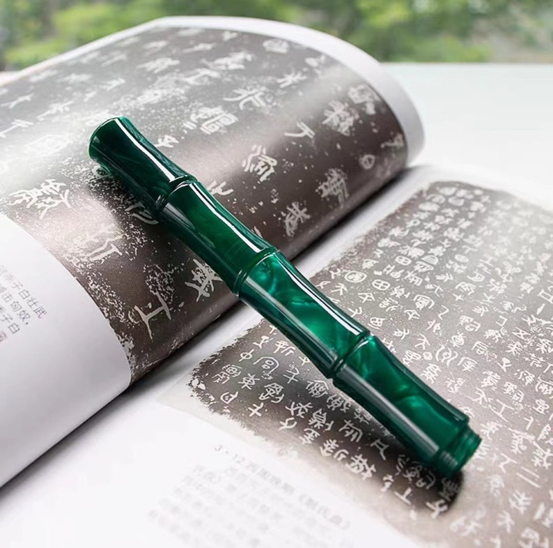 May include: A green fountain pen with a bamboo-like design. The pen is lying on a book with Chinese characters printed on the pages.