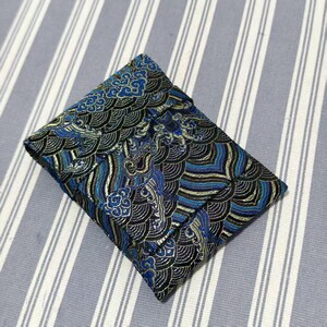 May include: A small, square pouch with a detailed wave pattern in shades of blue, black, and gold. The fabric has a textured appearance, and the pouch is closed. The background is a light blue and white striped fabric.