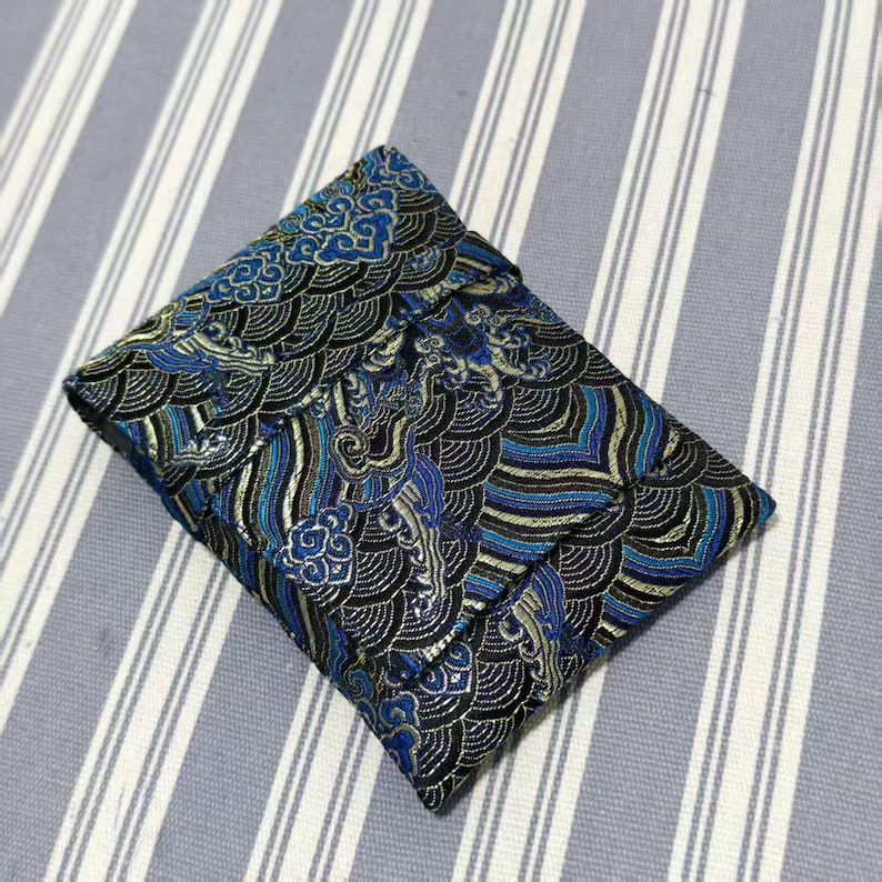 May include: A small, square pouch with a detailed wave pattern in shades of blue, black, and gold. The fabric has a textured appearance, and the pouch is closed. The background is a light blue and white striped fabric.