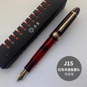 May include: A red and gold fountain pen with a clear body and a gold nib. The pen is in a black box with a red and white pattern. The box has the text "J15". The text "红色半透明鱼雷头" and "钛金笔" is also visible in the image.