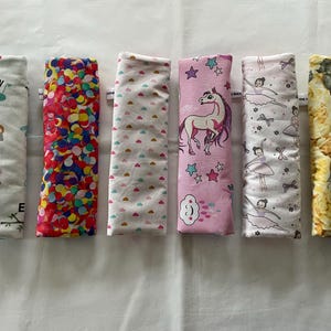 Könnte beinhalten: Sechs bunte Stoffbeutel mit verschiedenen Mustern. Die Beutel sind alle gleich groß und haben die gleiche Form und sind in einer Reihe angeordnet. Die Muster umfassen einen Regenbogen aus Konfetti, einen weißen Hintergrund mit rosa Herzen, einen rosa Hintergrund mit Sternen und einem Einhorn, einen weißen Hintergrund mit Ballerinas und einen gelben Hintergrund mit einem Katzenmuster.