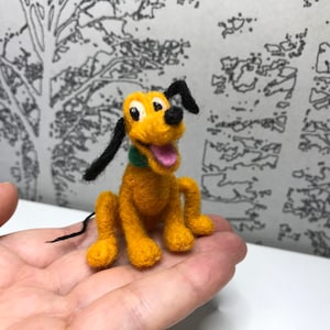 Può includere: Una piccola figurina di feltro gialla di Pluto, un cane dei cartoni animati Disney. Pluto è seduto con la lingua di fuori e le orecchie dritte.
