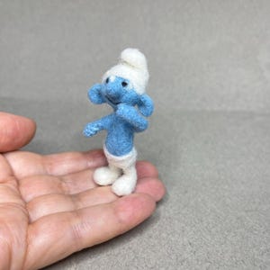 May include: A handmade felt Smurf figurine, featuring a blue body, white hat, and white pants. The miniature character is posed with arms outstretched, showcasing its whimsical design. The figure is approximately 2 inches tall.