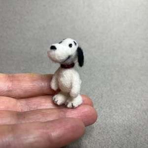 May include: A miniature needle-felted dog figurine crafted from white wool with a black ear and black spots on its head. The small animal figure features a dark red collar around its neck and has a rounded snout with a black nose and eyes.