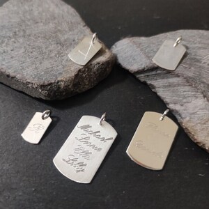 Könnte beinhalten: Fünf silberfarbene Erkennungsmarken-Anhänger aus Metall in verschiedenen Größen, jeweils mit einem kleinen Biegering an der Oberseite. Die Anhänger sind mit verschiedenen Namen und Buchstaben graviert, darunter Michael, Leonie, Ella und Libby in Schreibschrift.