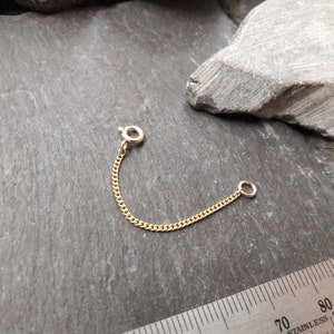 Könnte beinhalten: Ein goldener Kettenverlängerer mit einem Karabinerverschluss an einem Ende und einem Sprungring am anderen Ende. Die Kette ist etwa 5 cm lang.