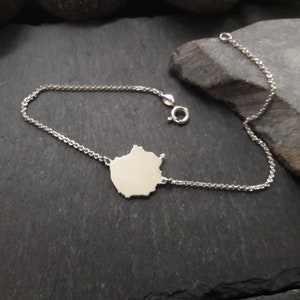 Könnte beinhalten: Ein silberfarbenes Kettenarmband mit einem mittigen, flachen, unregelmäßig geformten Anhänger mit glatter, reflektierender Oberfläche. Das Armband verfügt über einen kleinen runden Federringverschluss und ein feines Gliederkettendesign.